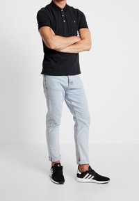 En person står med armarna i kors, iklädd en svart polo, ljusblå uppvikta jeans och svarta Adidas-sneakers med vita ränder.