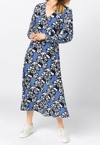 Dlouhý rukáv, překládané šaty v modré tkanině s černo-bílým květinovým vzorem, s výstřihem do V a volným siluetou, doplněné bílými teniskami.
