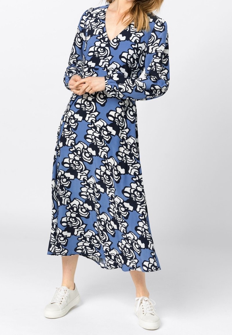 Dlouhý rukáv, překládané šaty v modré tkanině s černo-bílým květinovým vzorem, s výstřihem do V a volným siluetou, doplněné bílými teniskami.