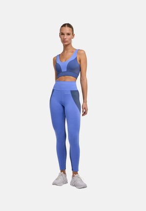 Frau steht und trägt einen nahtlosen Sport-BH und Leggings in Blau und Marineblau mit weißen Sneakers vor einfarbigem Hintergrund.