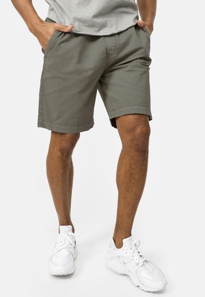 Mann som har på seg grå casual shorts med lommer, hvite treningssko, står med hendene i lommene mot en ensfarget bakgrunn.