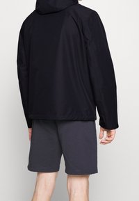 Person iført en sort hættejakke og mørkegrå knæ-lange shorts, set bagfra mod en ensfarvet lys baggrund.