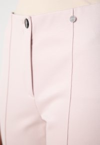 Pantalon rose clair sur mesure avec une fermeture à bouton noir et une petite étiquette ronde argentée avec logo près de la ceinture.