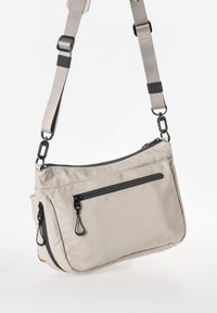 Beige Stoff-Schultertasche mit verstellbarem Gurt. Verfügt über schwarze Reißverschlüsse und Beschläge, zwei vordere Reißverschlussfächer und eine strukturierte Oberfläche.