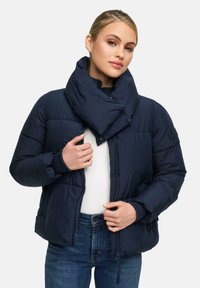 Vrouw in een marineblauwe gewatteerde jas met hoge kraag, die deze opent boven een witte top en blauwe jeans tegen een effen achtergrond.