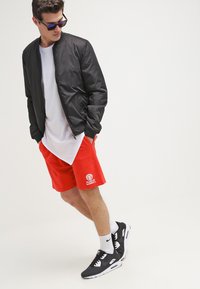 Chaqueta bomber negra, camiseta blanca, pantalones cortos rojos con logo, zapatillas negras, calcetines blancos y gafas de sol. Conjunto atlético casual con colores contrastantes.