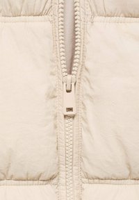 Nahaufnahme einer beigen Pufferjacke mit einem zentralen Reißverschluss, einem strukturierten Zugband, einem auffälligen gesteppte Muster und einer glatten Stoffoberfläche.