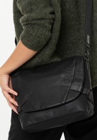 Black messenger bag made of textured fabric, featuring a fold-over flap, adjustable strap, and embossed logo on the front.