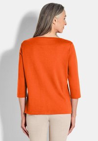 Rostorange Oberteil mit drei Viertel Ärmeln, geradem Schnitt und weitem Ausschnitt. Glatte Textur, minimale Nähte und keine sichtbaren Muster.