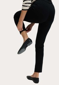 Pantalones ajustados negros con un dobladillo recortado, combinados con unas elegantes bailarinas negras. La mano del modelo ajusta la pierna del pantalón mientras levanta un pie.