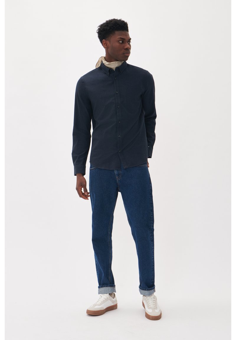 Camicia blu navy con colletto, abbinata a jeans blu arrotolati e sneakers bianche. La camicia presenta un tessuto testurizzato e dettagli di design discreti.