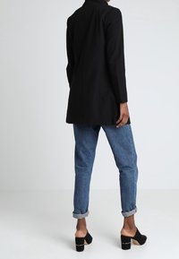 Blazer negro largo con un corte entallado, combinado con jeans azules remangados y mules negros de tacón abierto. Textura de tela suave.