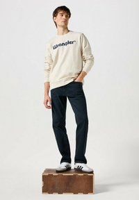 Felpa beige con logo "Wrangler", jeans blu scuro e scarpe bianche con strisce nere, in piedi su una scatola di legno.