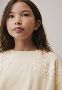 Světlý žlutý svetr s duhovými flitrovými akcenty tvořícími kaskádový vzor na přední straně. Měkká, texturovaná tkanina s kulatým výstřihem.