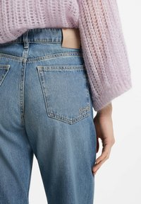 Jeans in denim azzurro chiaro con tasche posteriori e etichetta in pelle marrone chiaro in vita. Un maglione leggero, trasparente e lavorato a maglia color lavanda è parzialmente visibile.