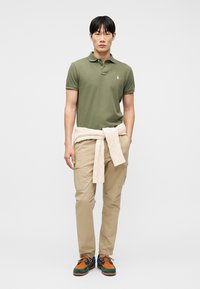 Νέος άνδρας που φοράει ένα χακί πράσινο πόλο, μπεζ παντελόνι cargo, μια κρεμ ζακέτα δεμένη γύρω από τη μέση του και πολύχρωμα παπούτσια με κορδόνια.