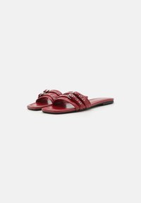 Red leather slide sandals with three horizontal straps, featuring metal hardware accents and a flat sole. Smooth texture and minimal design.