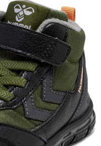 Zapatilla deportiva alta en negro y verde, con cuero y tejido texturizado, que presenta una correa de velcro, un collar acolchado y una tira de tiro naranja.