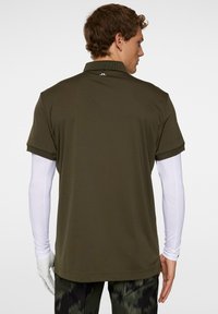 Polo shirt oliivinvihreä, pehmeällä tekstuurilla, lyhyillä hihoilla ja kauluksella. Valkoinen pitkähihaisen aluspaidan näkyy alla. Brändilogo takana.