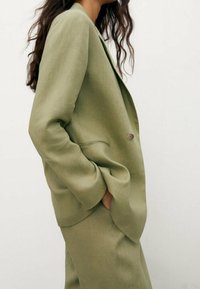 Olijfgroen linnen blazer met een ontspannen pasvorm, een enkele knoopsluiting en zijzakken, gecombineerd met bijpassende broek. Soepele textuur.