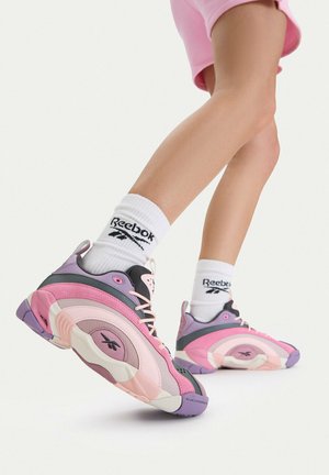 Beine, die pinkfarbene Shorts, weiße Reebok-Socken und pink-lila-schwarze Reebok-Sneaker tragen, vor einem weißen Hintergrund angehoben.