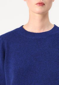 Pull en tricot bleu avec un col rond, texture douce et poignets côtelés. Le tissu a un aspect uniforme avec un éclat subtil.
