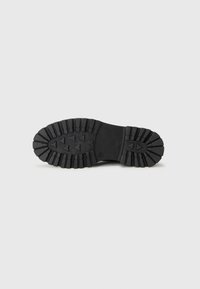 Black rubber shoe sole featuring a rugged tread pattern, with pronounced lugs for grip and a smooth central area for stability.