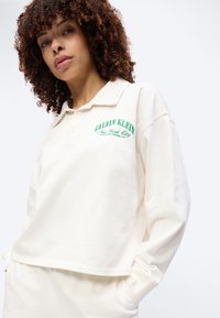 Γυναίκα με σγουρά μαλλιά που φοράει μια φαρδιά μακρυμάνικη πουλόβερ πόλο Calvin Klein σε κρεμ χρώμα και ταιριαστό παντελόνι, στέκεται με τα χέρια στις τσέπες.