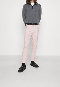 Grau zipfeliger Pullover, pinkfarbene Slim-Fit-Hosen, schwarzer Gürtel, schwarze Slipper, getragen mit schwarzen Socken. Glattes Material mit strukturiertem Sitz.