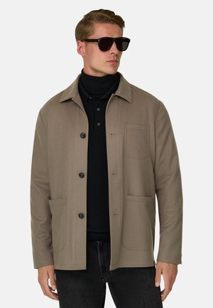 Homme portant des lunettes de soleil noires, une veste beige avec des boutons et des poches, un col roulé noir et un jean foncé, regardant vers la droite.