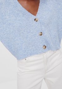 Mörkhyad person iklädd en ljusblå, fluffig knappad kofta och högmidjade off-white byxor mot en enfärgad bakgrund.