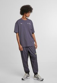 Pilkas oversized marškinėliai su Puma ir BMW logotipais, derinami su tamsiai pilkais sportiniais keliais su šviesiai žaliais akcentais, užsidėti su bėgimo bateliais.