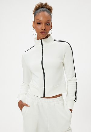 Witte korte ritsjack met hoge kraag, zwarte zijstrepen en een gestructureerde afwerking, gecombineerd met bijpassende sweatpants.