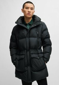 Black puffer coat with a zippered front, side pockets, and a high collar. Made from quilted fabric with a smooth texture. Button accents visible.