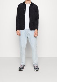 Svart kordflorsjacka med två bröstfickor och knappstängning, i kombination med ljusblå denimjeans och flerfärgade sneakers.