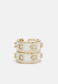 Brazalete de cuff de metal dorado con un acabado de esmalte blanco, que presenta logotipos circulares en relieve y acentos suaves en un diseño de doble banda.