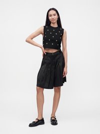 Femme debout portant un crop top noir sans manches avec des œillets dorés, un short noir plissé à hauteur des genoux, et des chaussures Mary Jane noires.