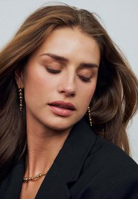 Guldkulor hänger från en modells öron. Hon har på sig en svart jacka och ett matchande svart halsband med rundade kulor. Håret flödar mjukt.