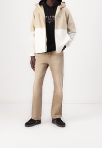 Kurtka wiatrówka w beżowo-białym kolorze z kapturem, zestawiona z czarną bluzą z kapturem i jasnobrązowymi prostymi dżinsami. Całość dopełniają czarne sneakersy.