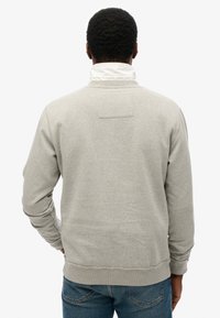 Sudadera gris de manga larga, con puños y dobladillo acanalados, que presenta un pequeño parche rectangular en la parte posterior. Lleva con jeans azules.