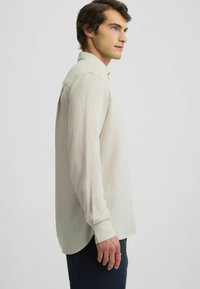 Camicia beige chiaro a maniche lunghe in tessuto morbido, con colletto classico, polsini abbottonati e orlo curvo, indossata sopra pantaloni blu navy.