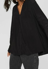 Blouse noire à manches longues en tissu léger, avec un décolleté en V, une fermeture à boutons à l'avant et une coupe légèrement détendue avec un détail plissé aux épaules.