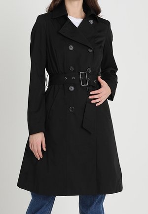 Femme portant un trench-coat noir croisé avec ceinture et boucle, associé à un jean bleu, debout avec une main sur le manteau.