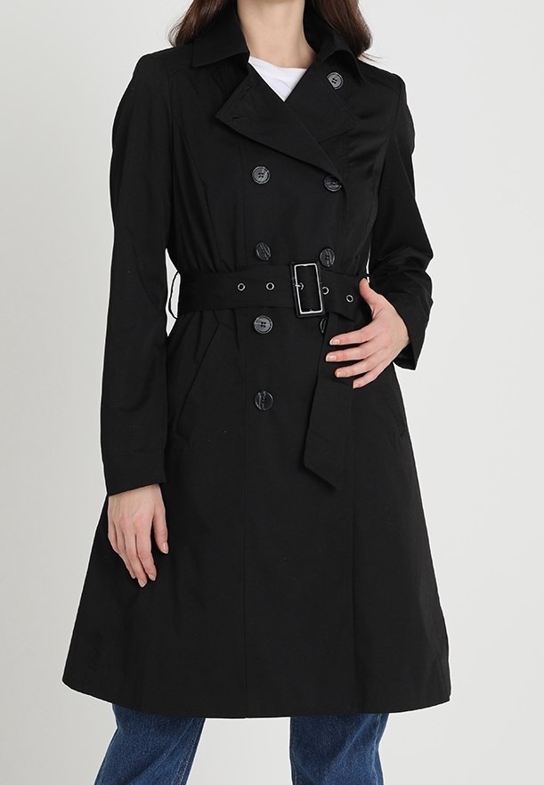 Femme portant un trench-coat noir croisé avec ceinture et boucle, associé à un jean bleu, debout avec une main sur le manteau.