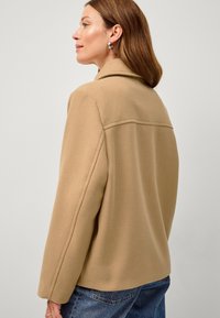Beige uld-blandet jakke med klapkrave og lange ærmer. Har en søm på tværs af ryggen, parret med denimjeans. Glat tekstur.