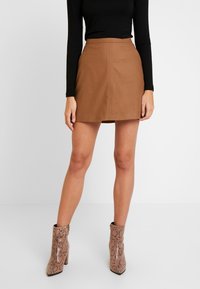 Femme portant une mini-jupe en suède couleur camel, un haut noir à manches longues et des bottines à motif serpent, se tenant contre un fond blanc.