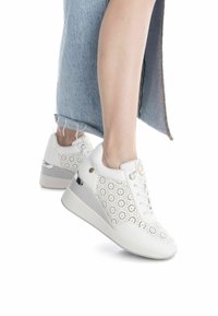 Zapatillas blancas con un diseño floral perforado, suela blanca plana y sutiles acentos metálicos. El zapato tiene una textura de cuero suave.