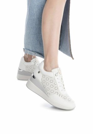 Zapatillas blancas con un diseño floral perforado, suela blanca plana y sutiles acentos metálicos. El zapato tiene una textura de cuero suave.
