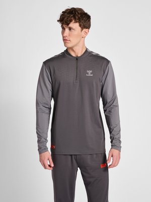 Jeune homme portant un haut de sport gris foncé à manches longues et un pantalon assorti, regardant vers sa gauche sur un fond clair uni.