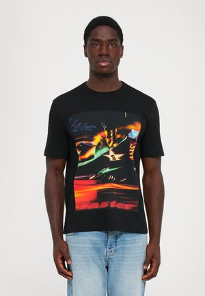 Man wearing black t-shirt with colorful abstract graphic and light blue jeans, standing against a plain white background.